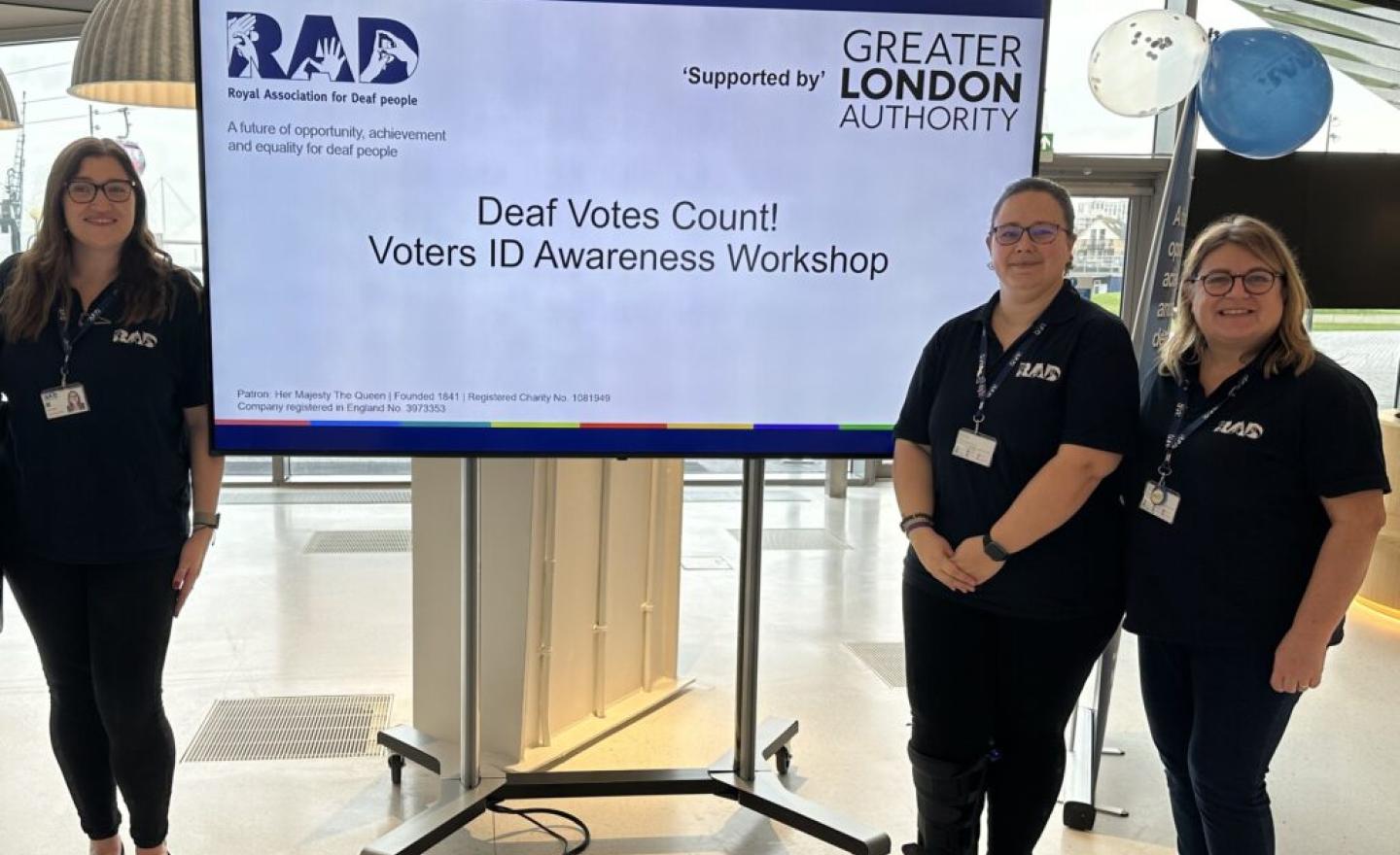 Three RAD staff standing in front of a large screens that displays the words 'Deaf Votes Count'