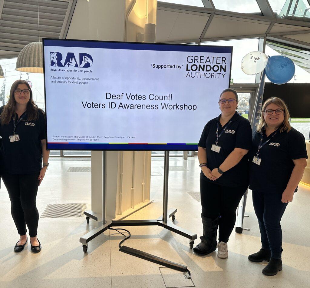Three RAD staff standing in front of a large screens that displays the words 'Deaf Votes Count'